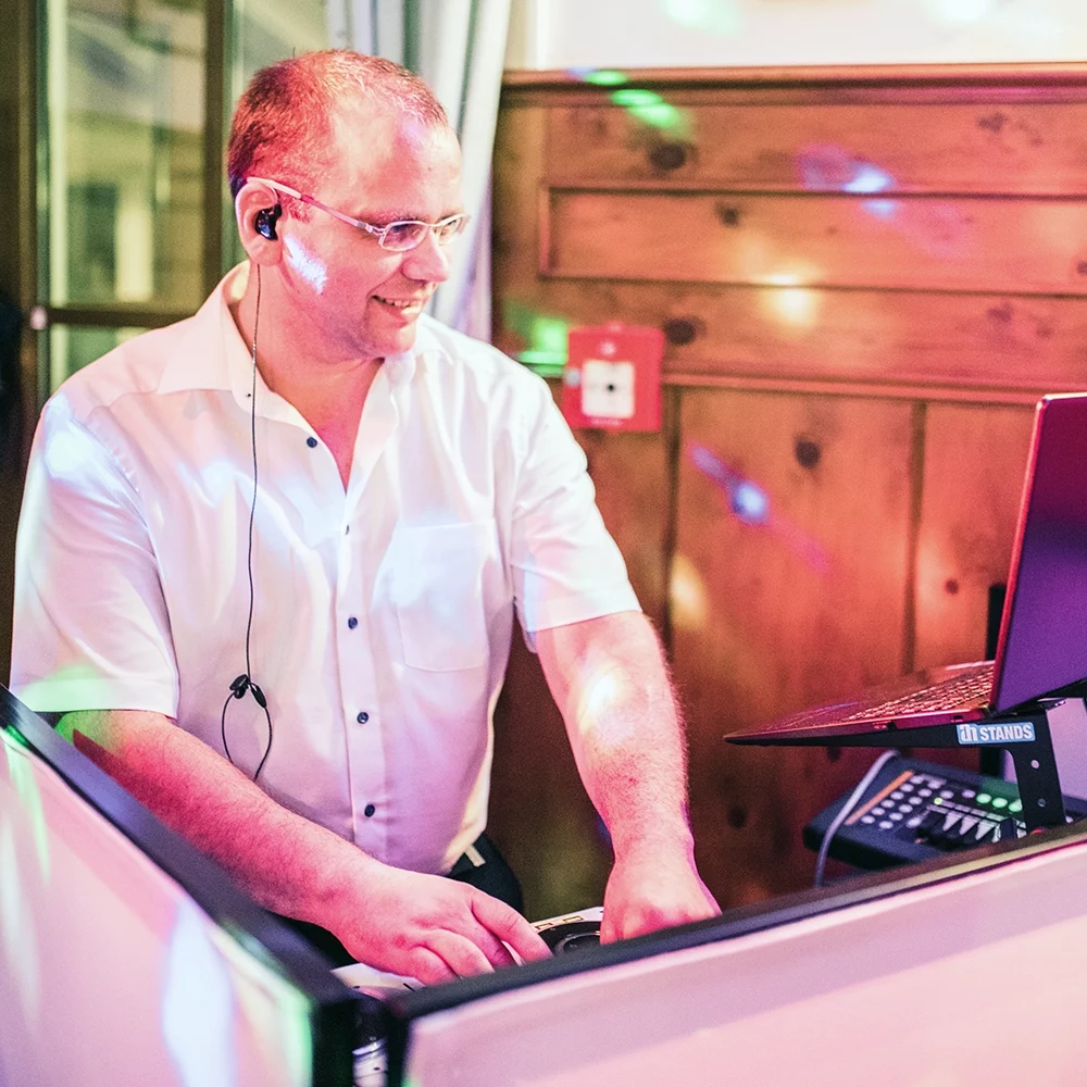 DJ mit kurzen braunen Haaren und Brille in weißem kurzen Hemd steht hinter DJ Pult und schaut auf den Bild schirm eines Acer Notebooks, vor hölzerner Wand.