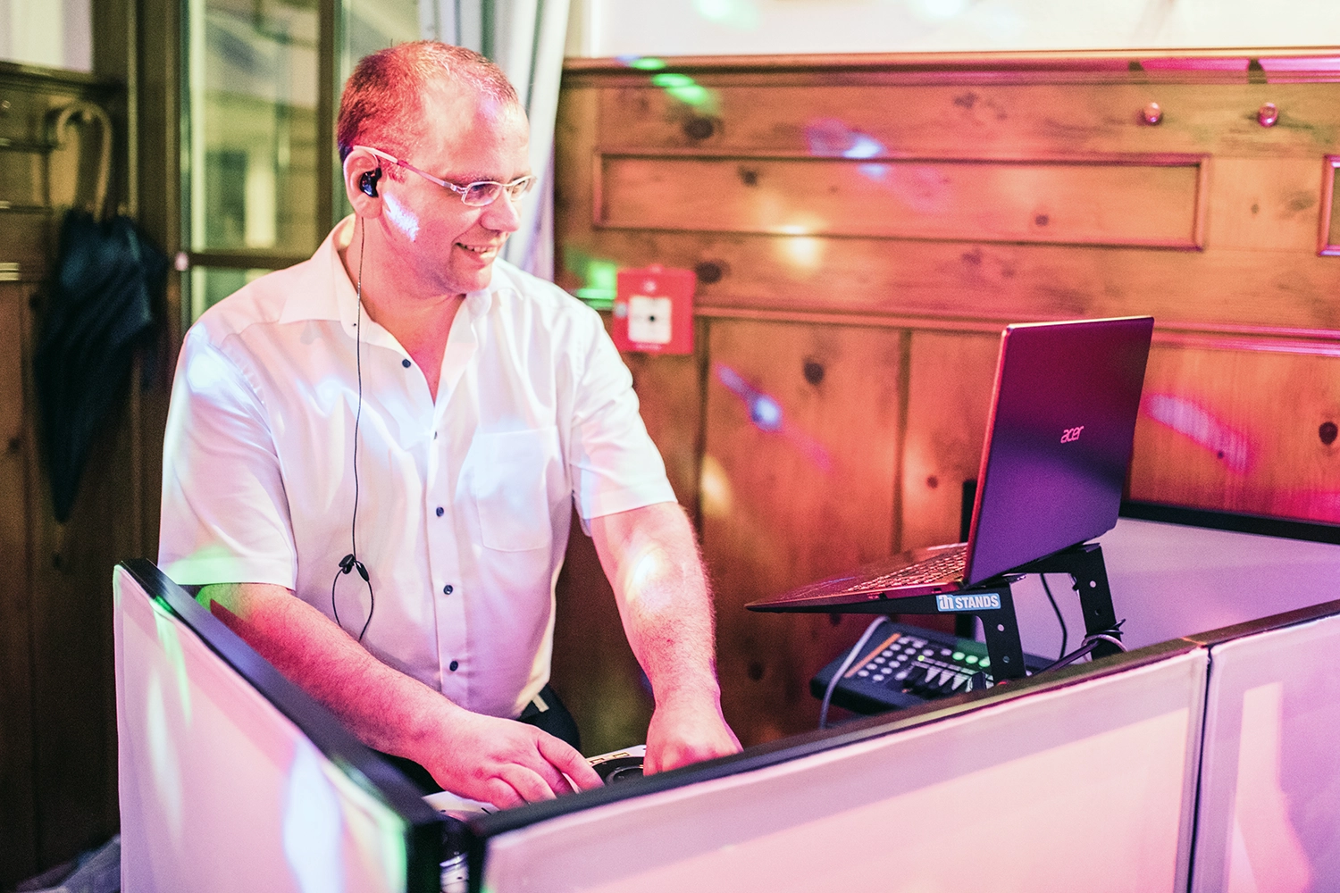 DJ mit kurzen braunen Haaren und Brille in weißem kurzen Hemd steht hinter DJ Pult und schaut auf den Bild schirm eines Acer Notebooks, vor hölzerner Wand.