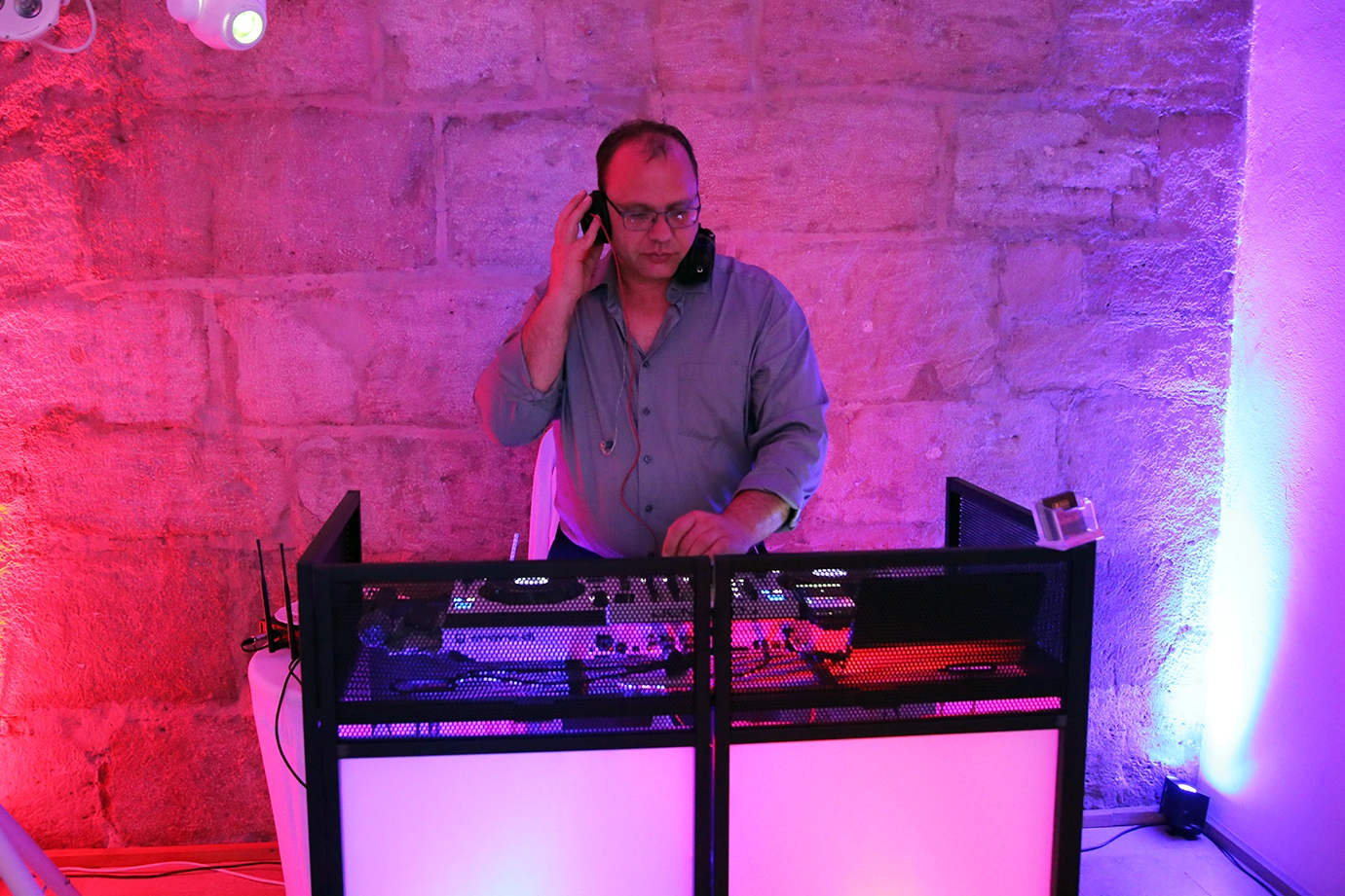 DJ mit kurzen braunen Haaren und Brille in grauem Hemd steht hinter DJ Pult vor einer pink beleuchteten Wand und hält sich mit einer Hand den Kopfhörer ans Ohr.
