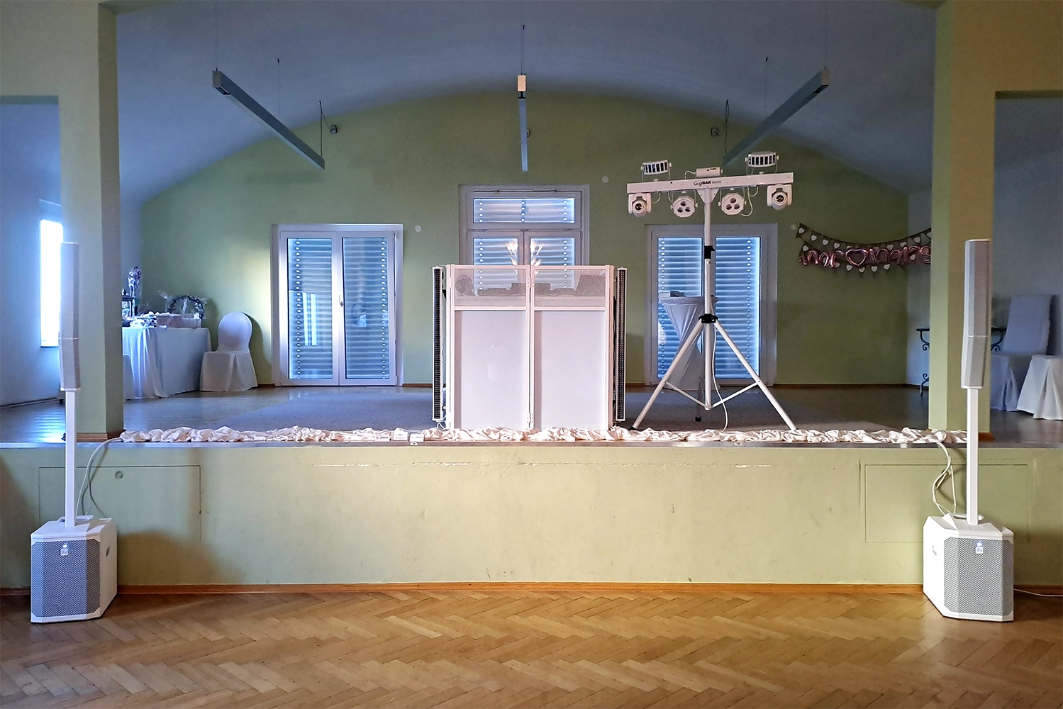 Elegante weiße DJ-Anlage steht auf hellgrüner Bühne mit drei Fenstern im Hintergrund.