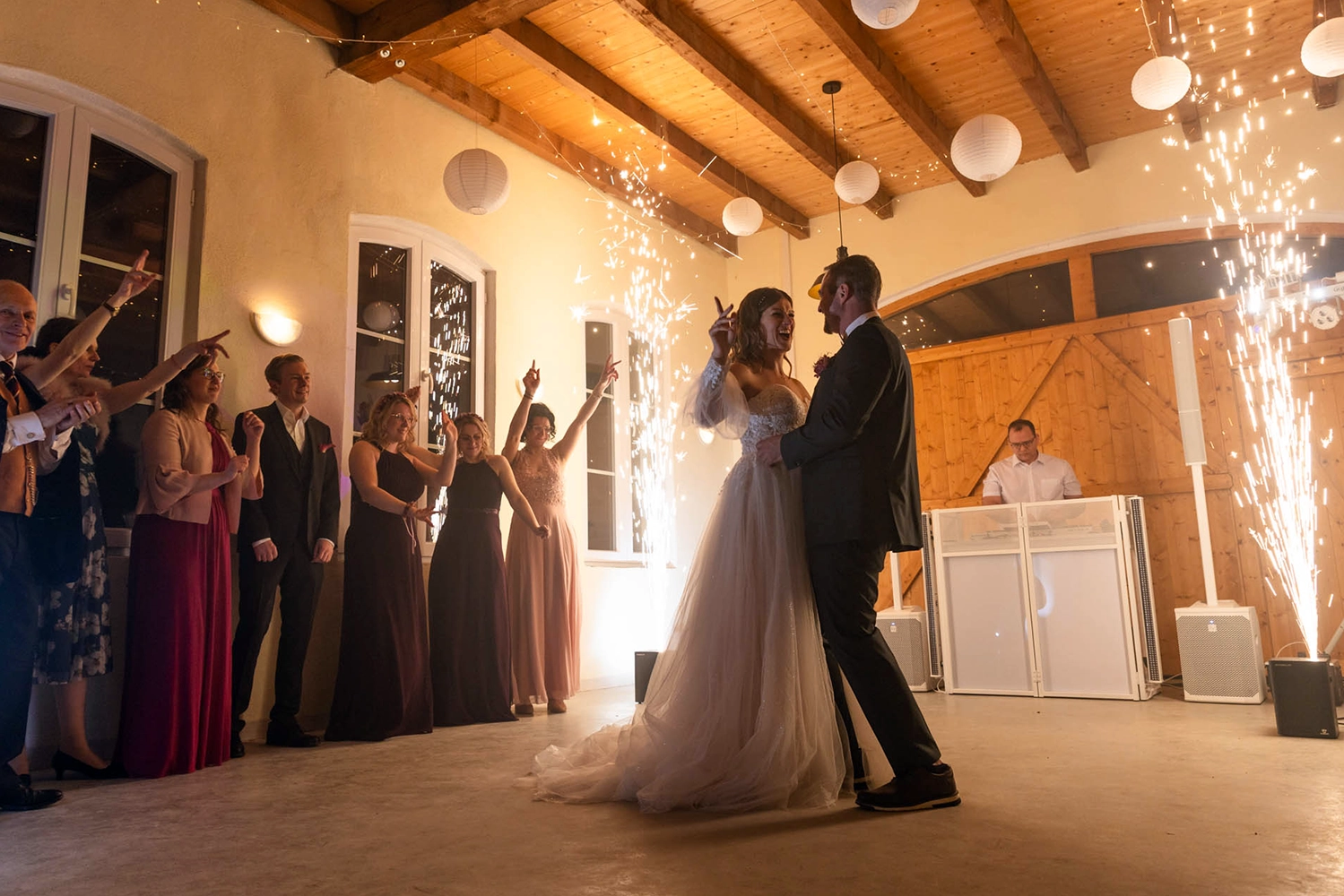 Tanzendes Brautpaar neben Hochzeitgesellschaft , während im Hintergrund Feuerwerks-Fontänen sprühen und DJ Jürgen Musik spielt.