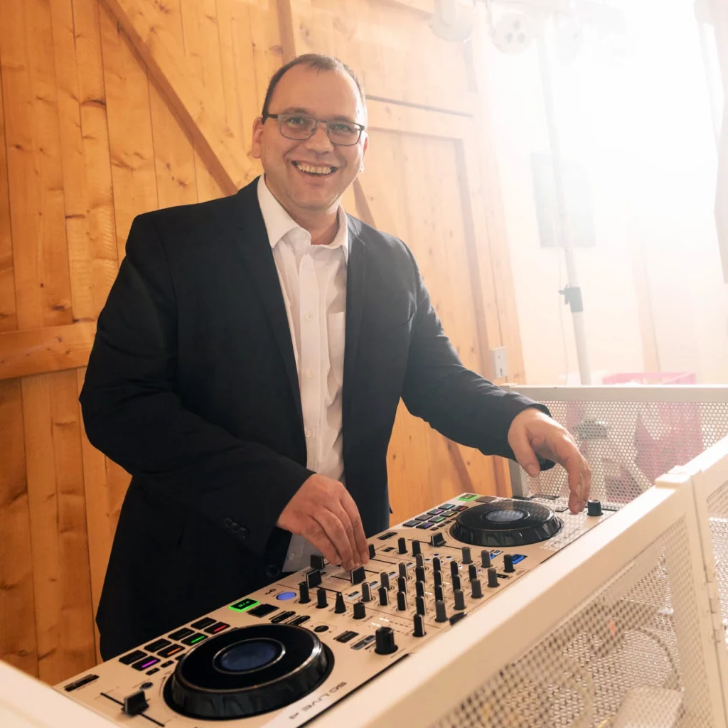Hochzeits-DJ Jürgen im Anzug steht lächelnd am DJ-Pult und mischt Musik vor einem hellen hölzernen Scheunentor.