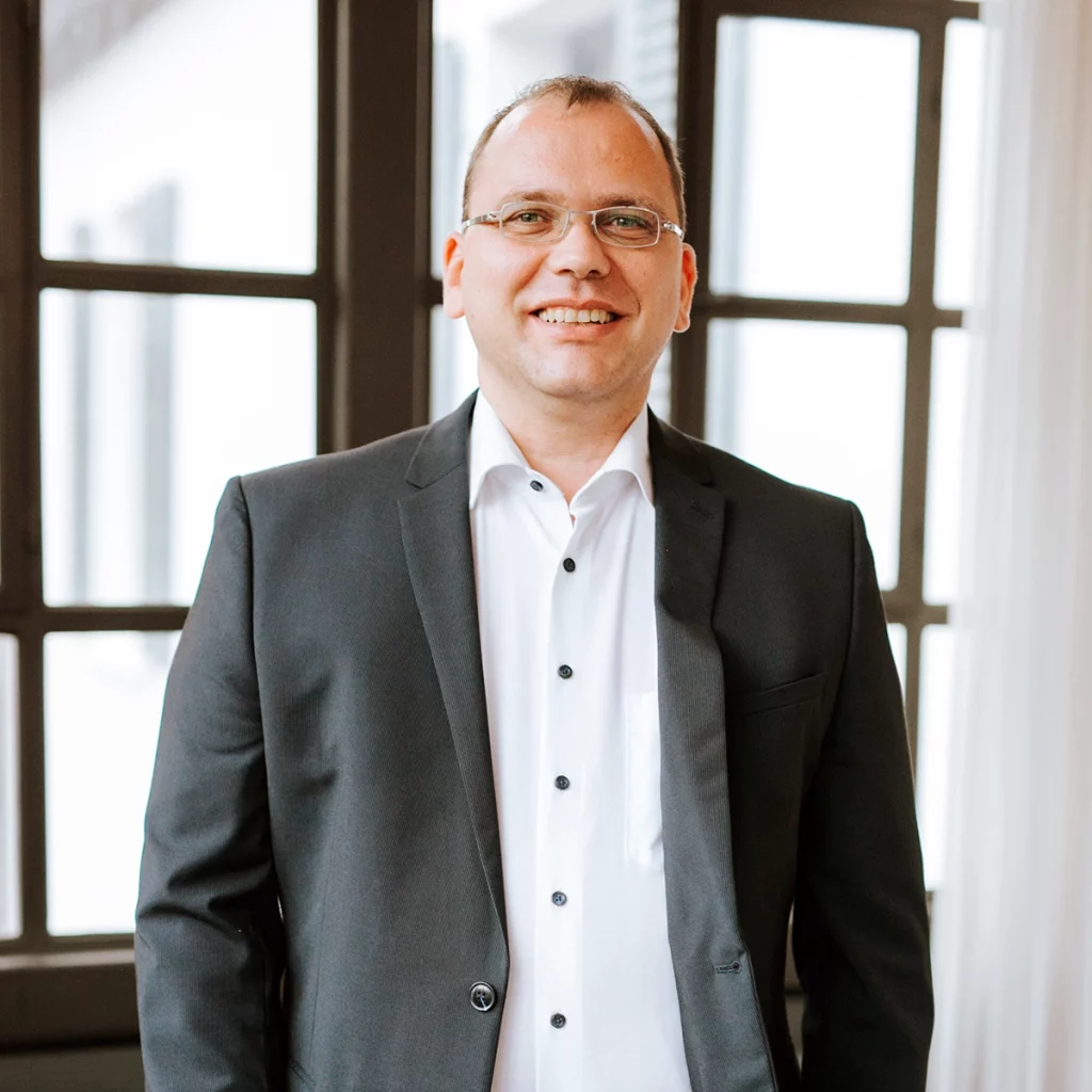 Mann mit kurzen braunen Haaren und Brille in weißem und schwarzem Anzug blickt lächelnd in die Kamera.